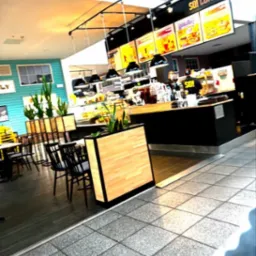 Kawiarnia z drewnianymi donicami i czarnymi lampami wiszącymi nad ladą. Turkusowa ściana w tle. Ustawienie stolików i krzeseł. Czarny blat.