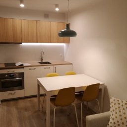 Aura Tek Sp. z o.o. - Nowoczesna kuchnia: drewniane szafki, białe fronty, stół z żółtymi krzesłami i designerska lampa. Parkiet na podłodze. Ukończona aranżacja wnętrza.