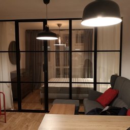 Aura Tek Sp. z o.o. - Nowoczesny salon z czarną, przeszkloną ścianą działową, oświetlony wiszącymi lampami. Widoczne meble: czerwona komoda, szara sofa i drewniany stół.