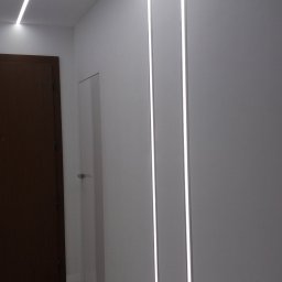 Aura Tek Sp. z o.o. - Nowoczesne oświetlenie LED w korytarzu: białe ściany, marmurowa podłoga, minimalistyczne listwy świetlne na ścianie i suficie. Widoczny fragment drzwi i narzędzia.