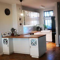 Aura Tek Sp. z o.o. - Jasna kuchnia z drewnianym blatem i podłogą w jodełkę. Białe szafki z przeszkleniami, czarny zegar na ścianie i designerska lampa wisząca.