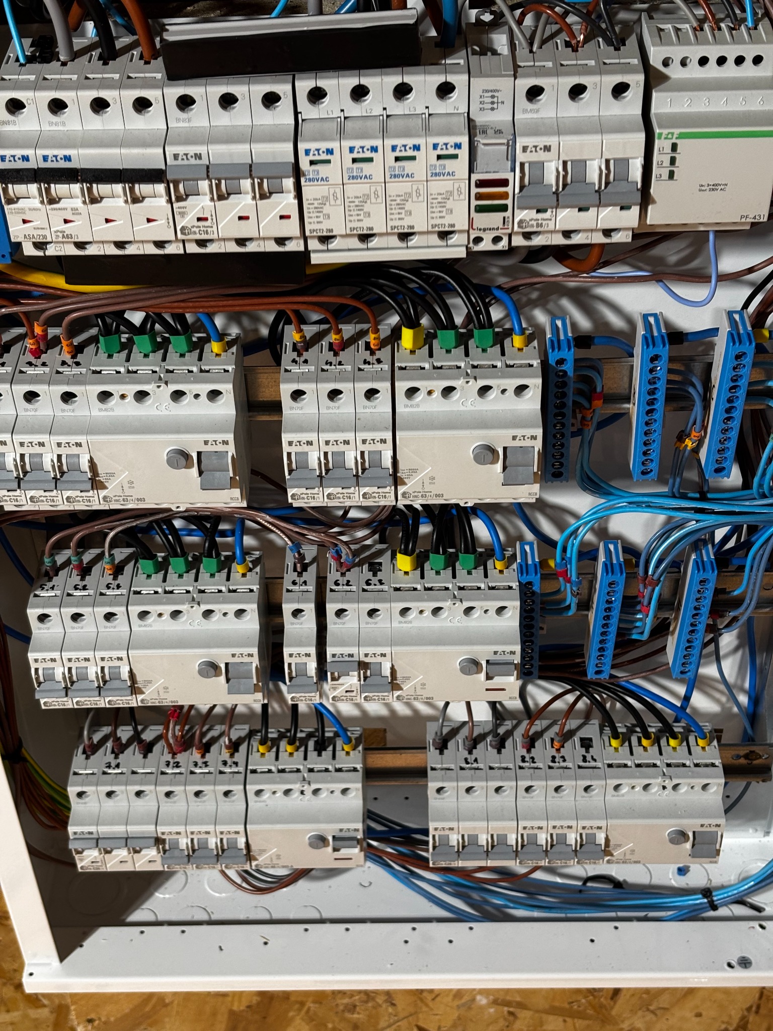 Rozdzielnica elektryczna z widocznymi bezpiecznikami EATON, przewodami w różnych kolorach (niebieski, czarny, brązowy, żółto-zielony) oraz listwami zaciskowymi. Widok na rozłożoną instalację...