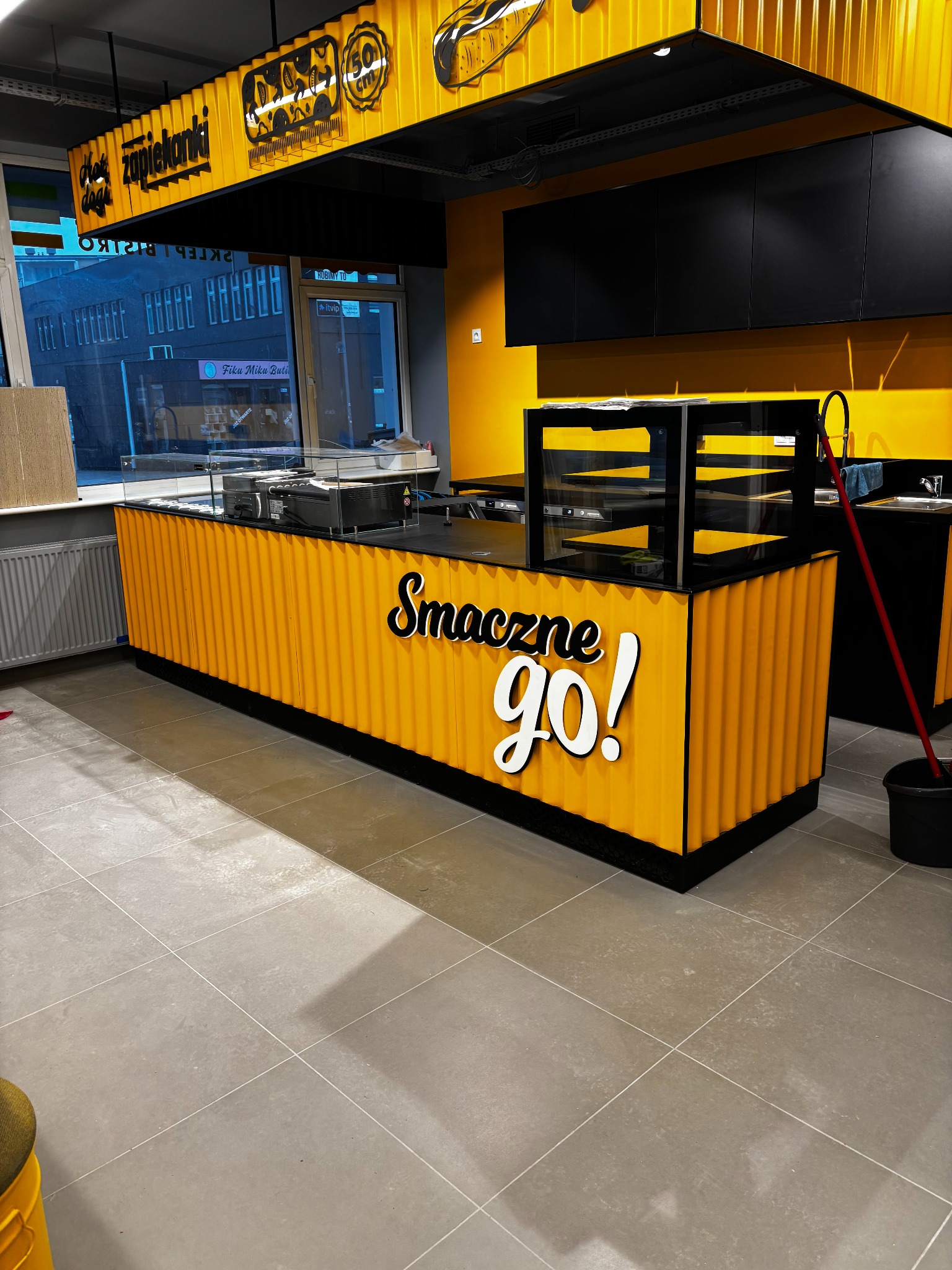 Wnętrze lokalu gastronomicznego w trakcie sprzątania: żółta lada z napisem 'Smaczne GO!', czarne szafki, mop i wiadro na szarej podłodze z płytek.