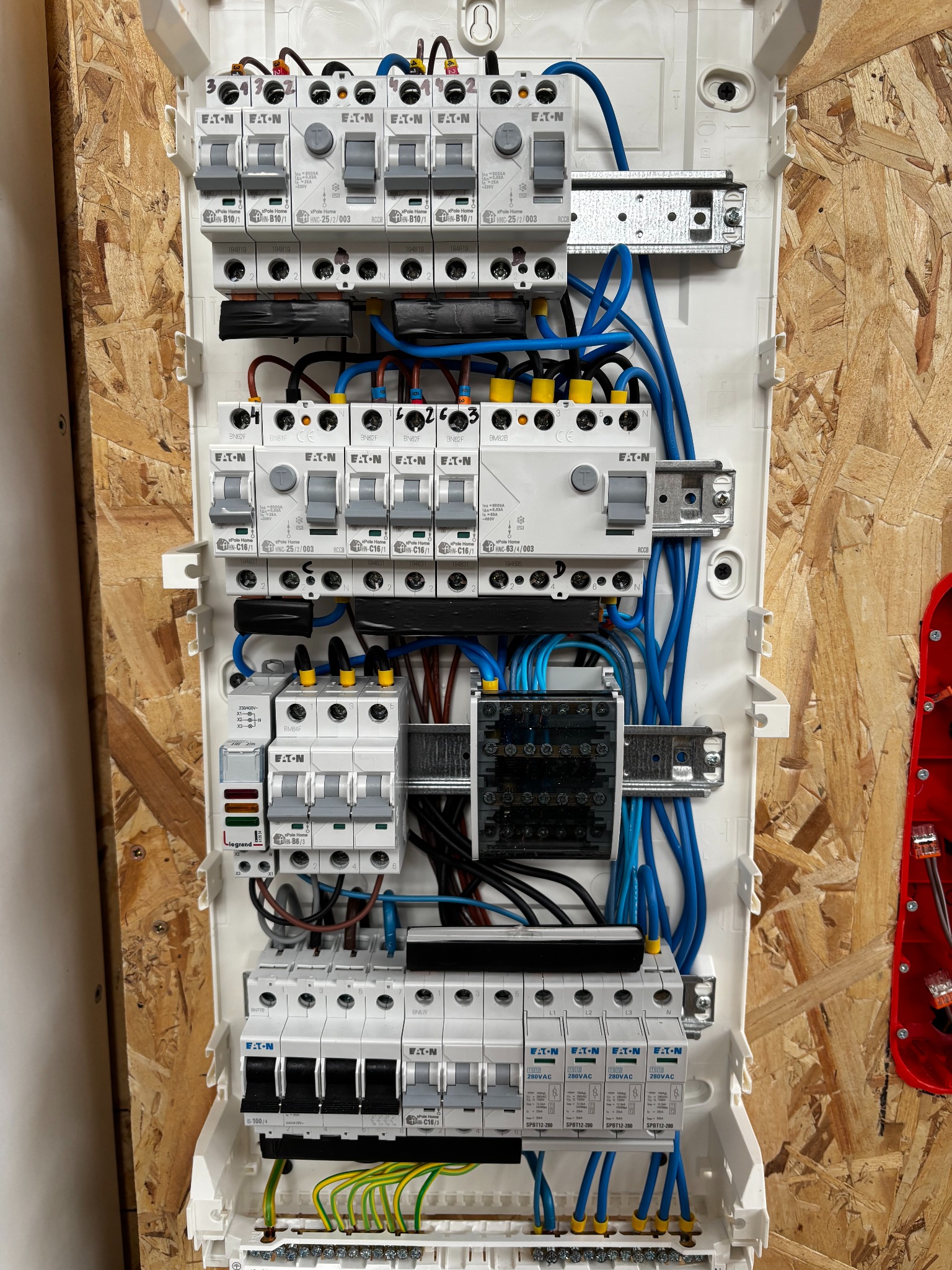 Rozdzielnica elektryczna z zainstalowanymi bezpiecznikami EATON, widoczne kable w kolorze niebieskim, brązowym i żółto-zielonym, zamontowana na płycie OSB.