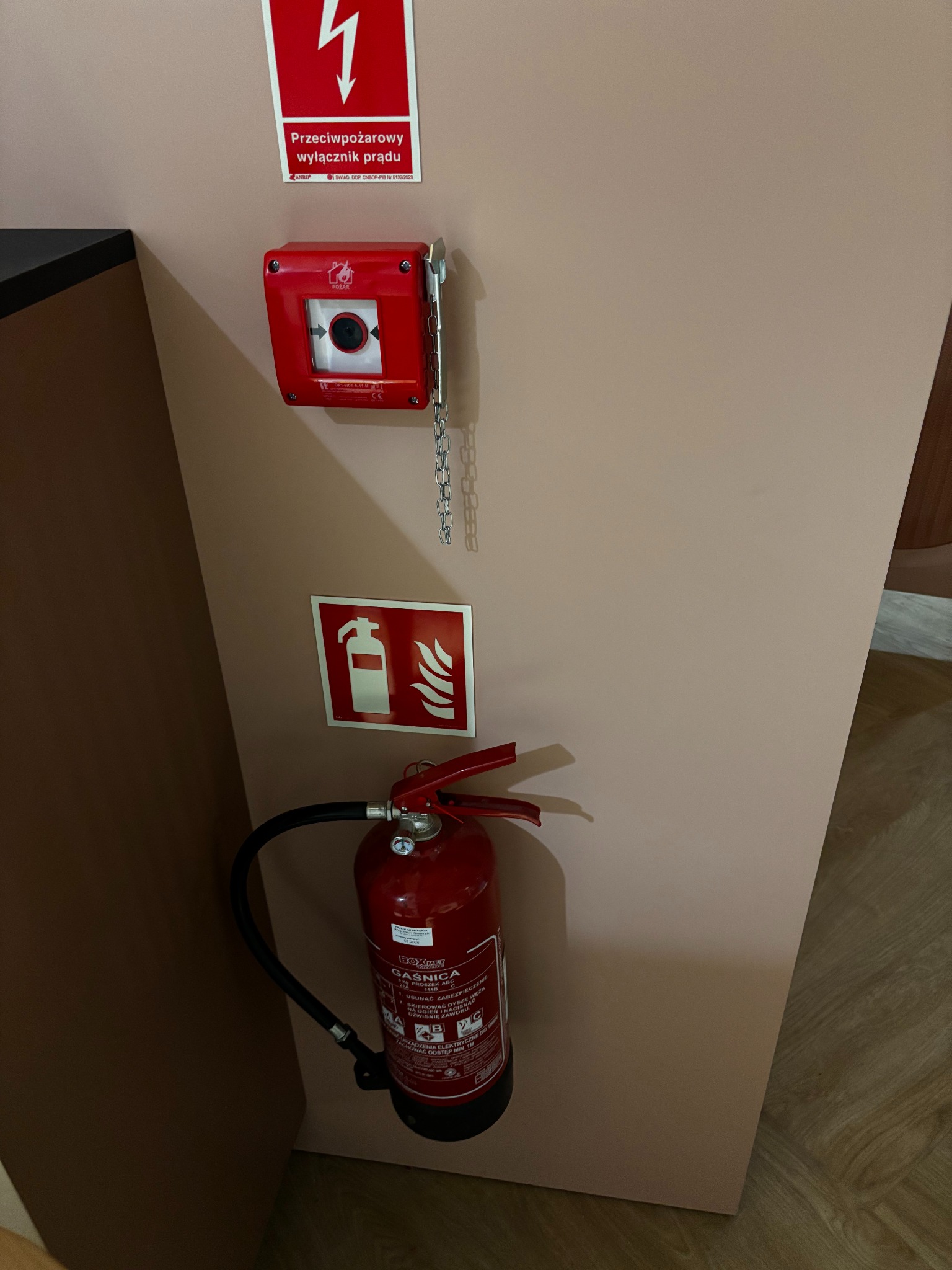 Czerwony wyłącznik prądu przeciwpożarowego z łańcuszkiem, gaśnica proszkowa i znak graficzny gaśnicy na ścianie w neutralnym kolorze.
