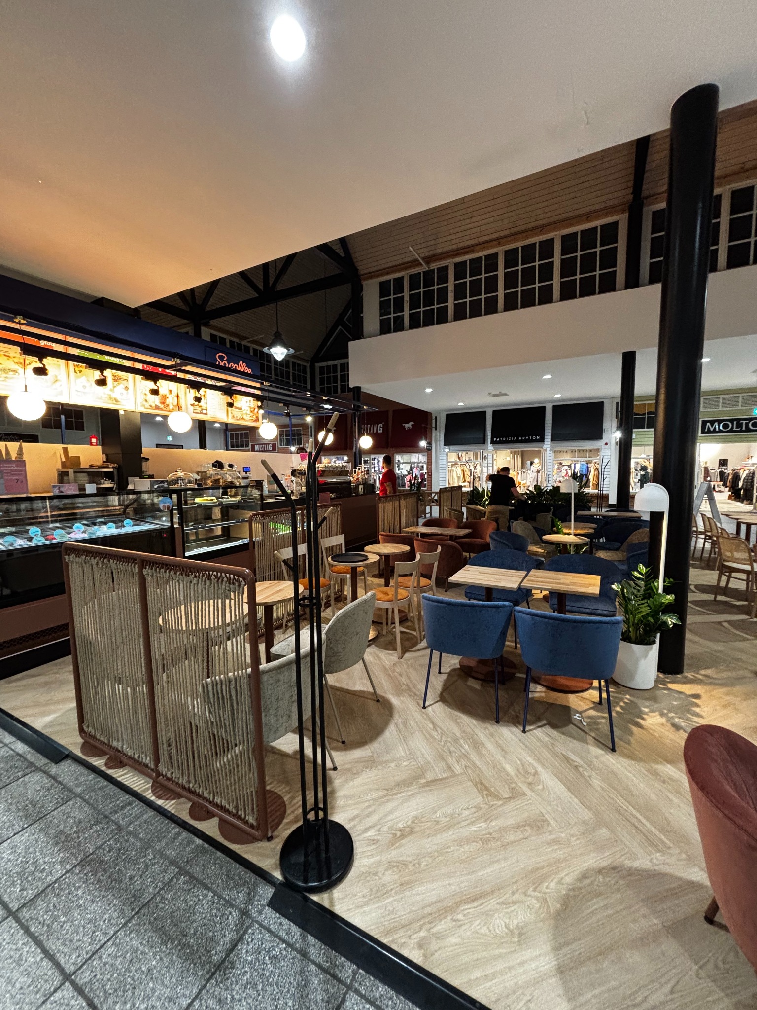 Wnętrze kawiarni z widocznym barem z ciastami i lodami, stolikami z niebieskimi krzesłami i roślinami w donicach, oświetlone jasnym światłem.