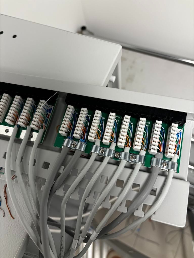 Panel krosowniczy z podłączonymi przewodami UTP, zamocowanymi metalowymi uchwytami. Widoczne kolorowe żyły w gniazdach i szare kable prowadzone w listwie.