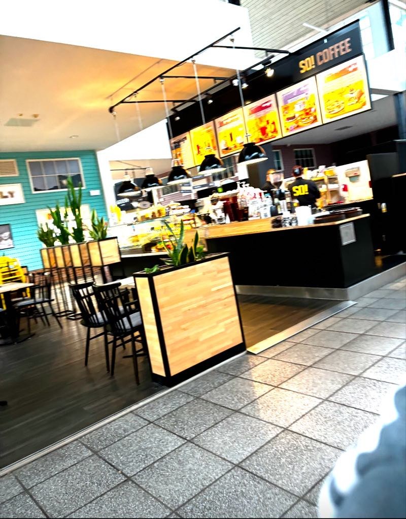 Kawiarnia z drewnianymi donicami i czarnymi lampami wiszącymi nad ladą. Turkusowa ściana w tle. Ustawienie stolików i krzeseł. Czarny blat.