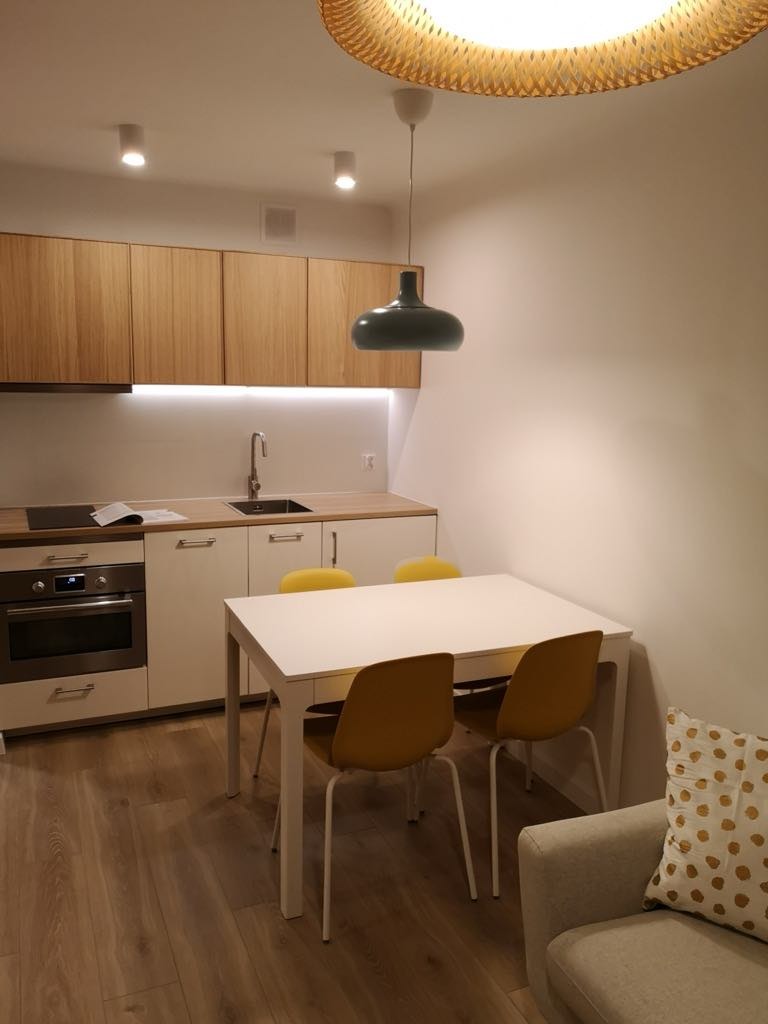 Nowoczesna kuchnia: drewniane szafki, białe fronty, stół z żółtymi krzesłami i designerska lampa. Parkiet na podłodze. Ukończona aranżacja wnętrza.