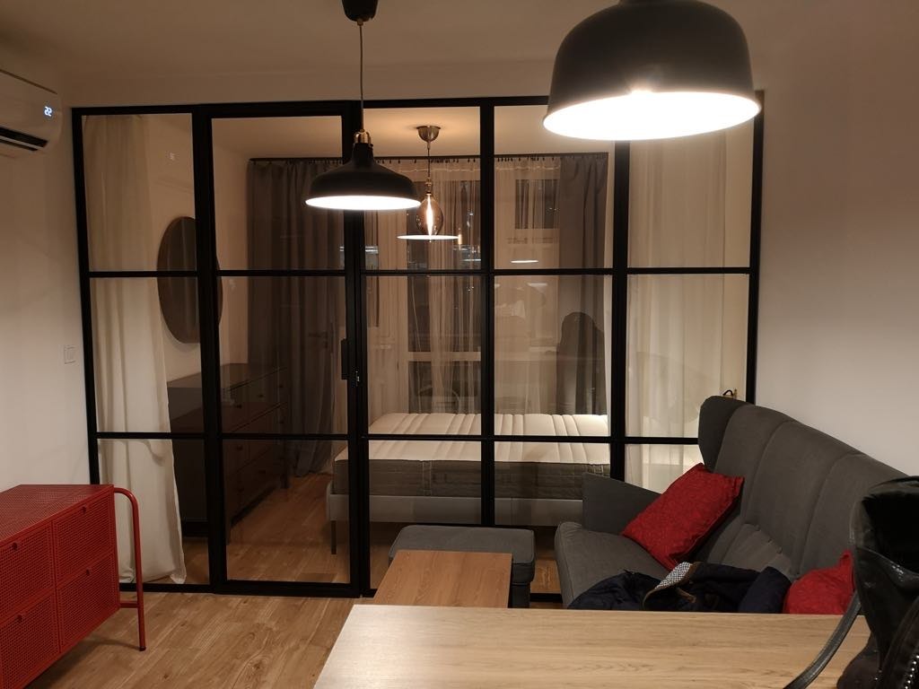 Nowoczesny salon z czarną, przeszkloną ścianą działową, oświetlony wiszącymi lampami. Widoczne meble: czerwona komoda, szara sofa i drewniany stół.