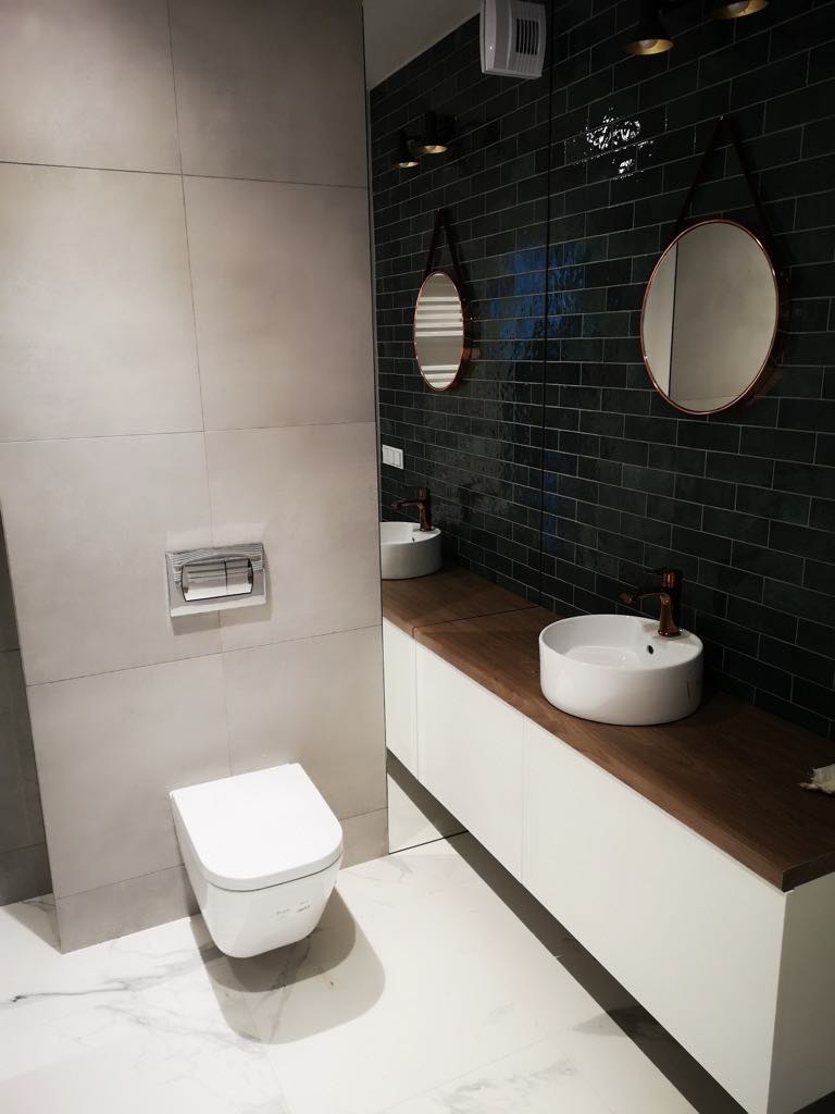 Nowoczesna łazienka z ciemnozielonymi płytkami, okrągłymi lustrami z miedzianymi ramami i białą ceramiką. Marmurowa podłoga z szarymi żyłami.