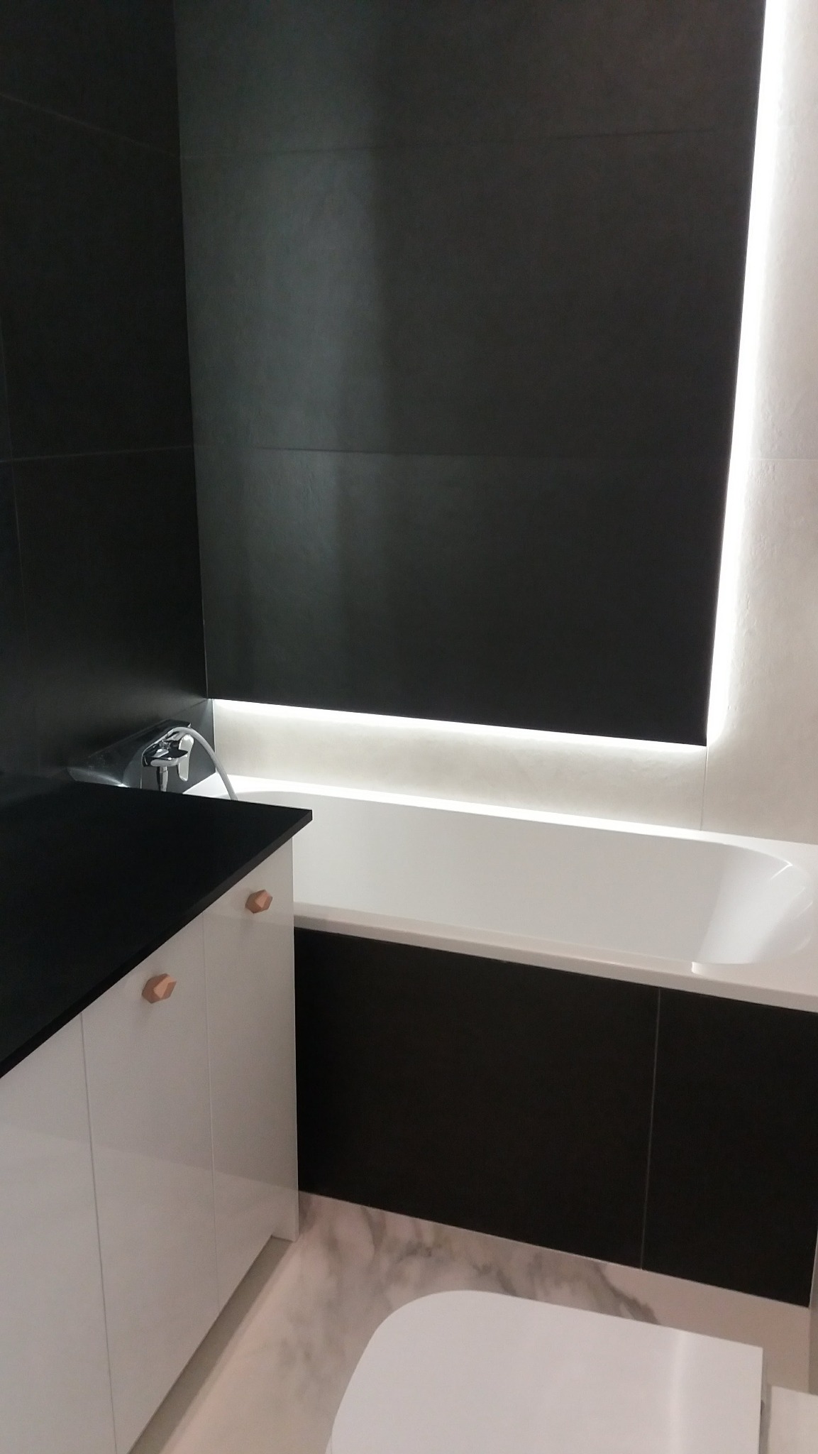 Nowoczesna łazienka z czarno-białymi płytkami, podświetlaną wanną i szafką z drewnianymi uchwytami. Minimalistyczny design, marmurowa podłoga.