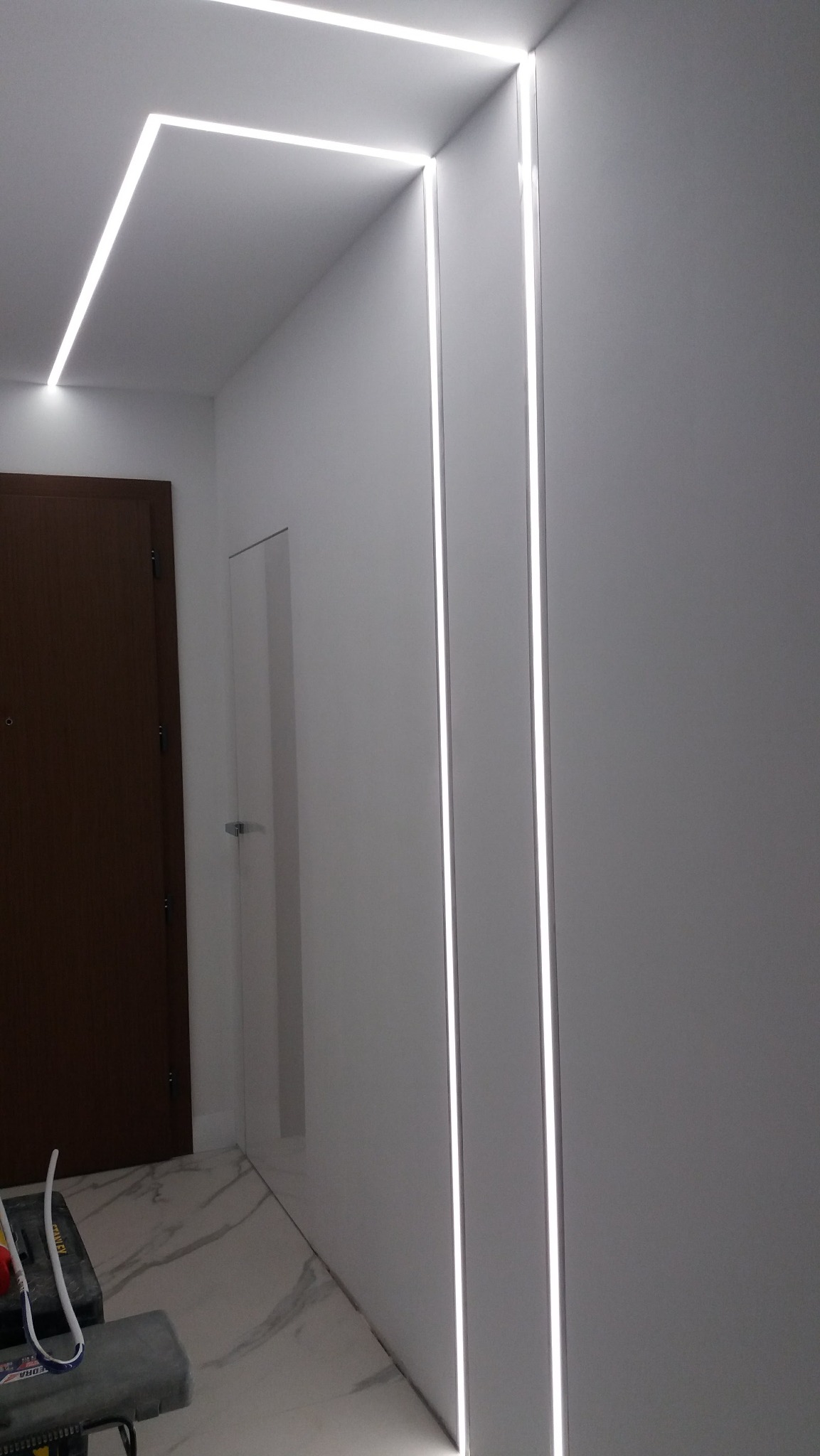 Nowoczesne oświetlenie LED w korytarzu: białe ściany, marmurowa podłoga, minimalistyczne listwy świetlne na ścianie i suficie. Widoczny fragment drzwi i narzędzia.