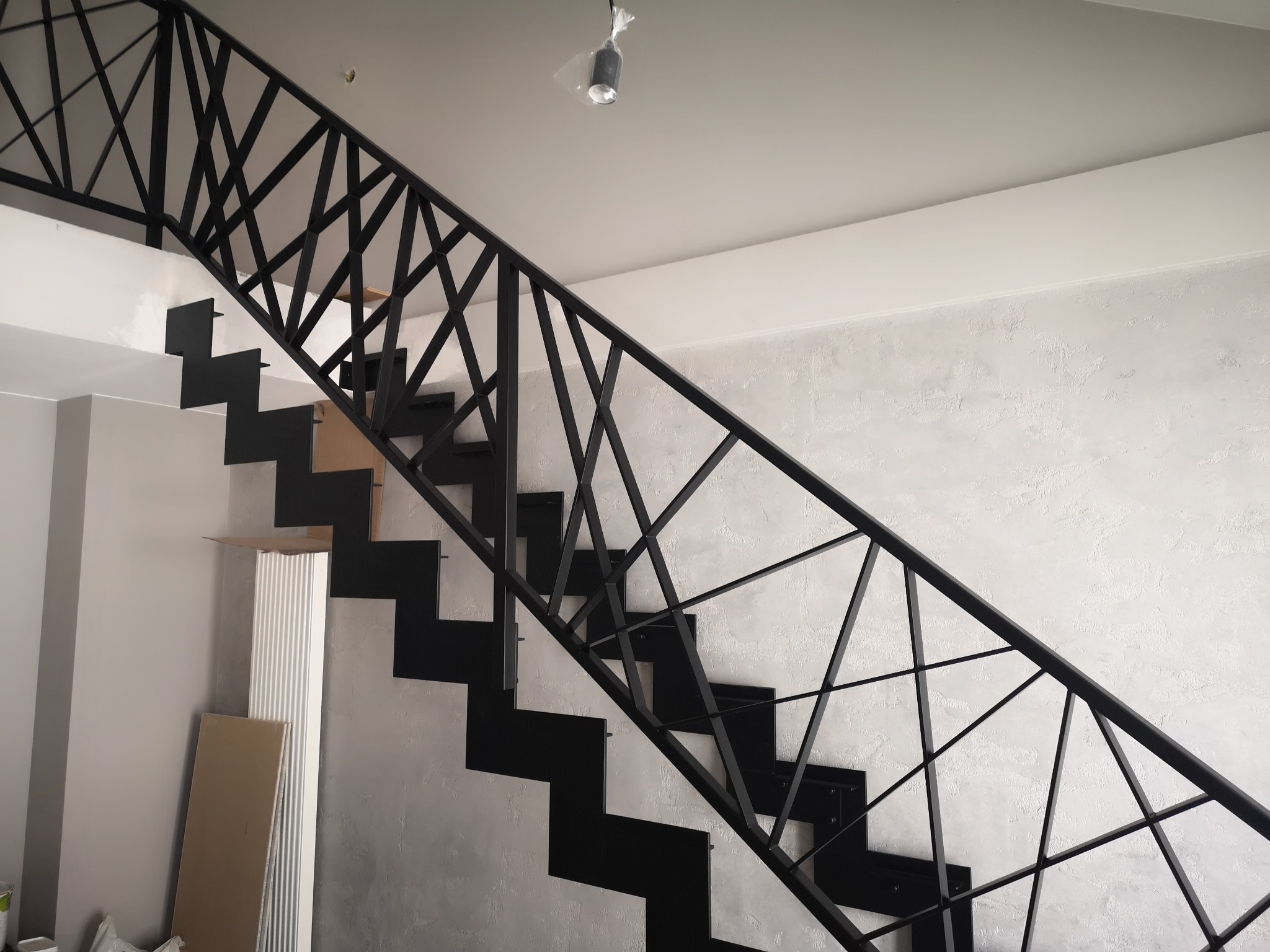 Nowoczesne, czarne schody z metalową balustradą o geometrycznym wzorze, na tle ściany o fakturze betonu. W tle widoczne materiały budowlane.
