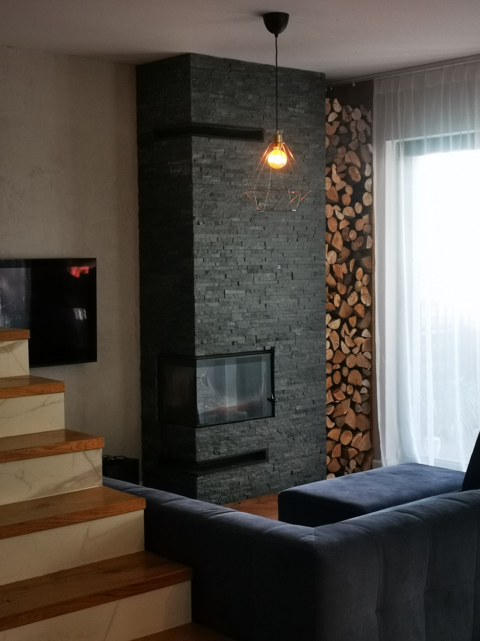 Nowoczesny salon z kominkiem obłożonym szarym kamieniem, drewnem obok okna i designerską lampą wiszącą. Widoczne schody z drewnianymi stopniami i fragment sofy.