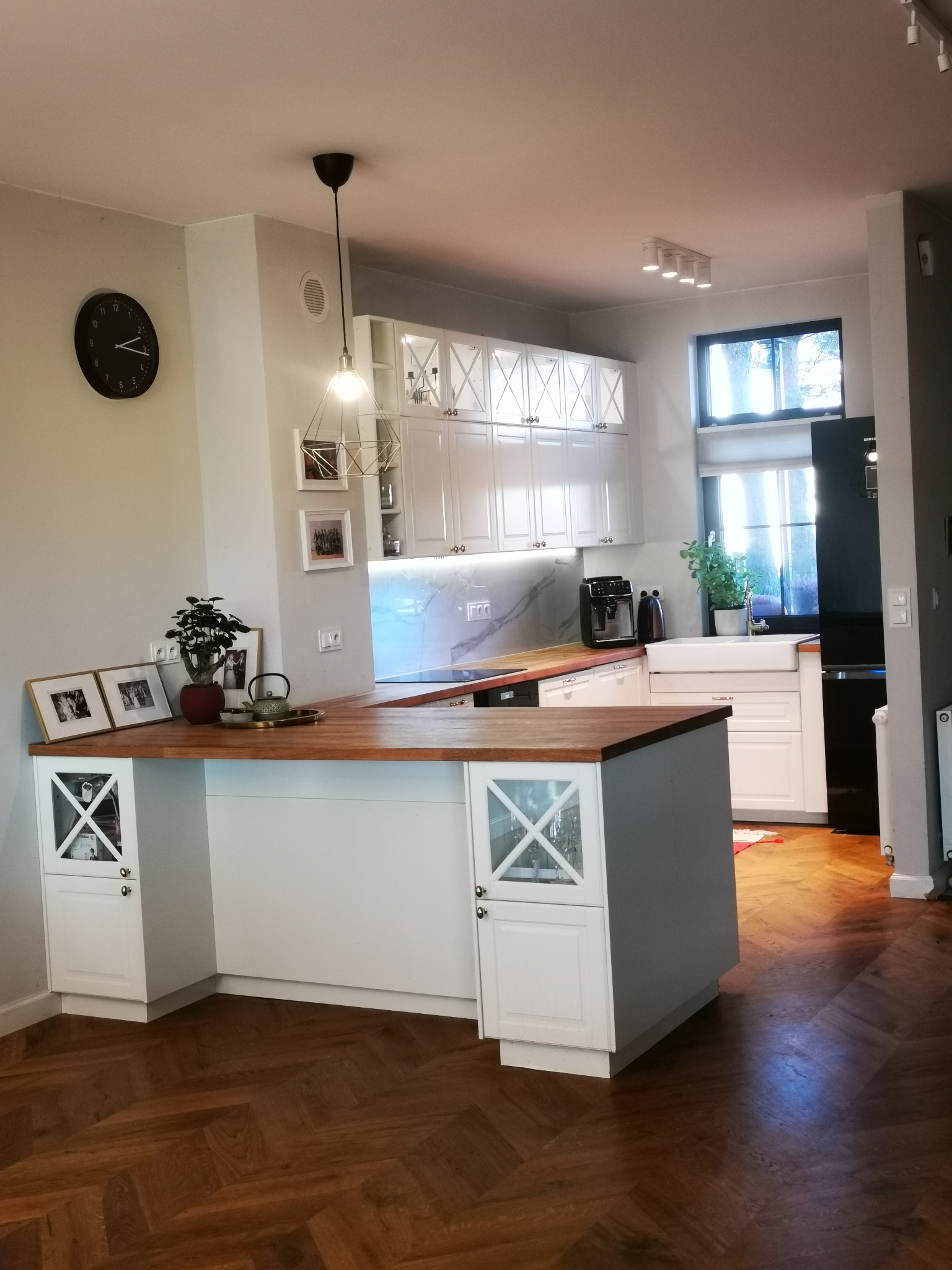 Jasna kuchnia z drewnianym blatem i podłogą w jodełkę. Białe szafki z przeszkleniami, czarny zegar na ścianie i designerska lampa wisząca.