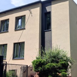 PrimeHome Oleksandr Prydvornyi - Dwukondygnacyjny dom z beżową elewacją, czarnymi ramami okiennymi i pionowym pasem w kolorze antracytowym, widoczny zza metalowego ogrodzenia i zieleni krzewów.