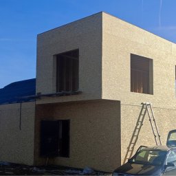 PrimeHome Oleksandr Prydvornyi - Dwukondygnacyjny dom w konstrukcji szkieletowej, z widocznym poszyciem z płyt OSB, częściowo pokryty dachem, z otworami okiennymi i drzwiami, drabina oparta o ścianę, otwarty bagażnik samochodu...