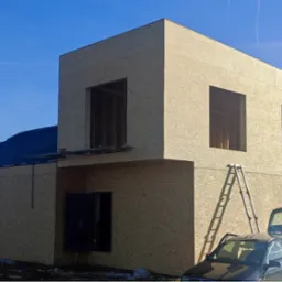 Dwukondygnacyjny dom w konstrukcji szkieletowej, z widocznym poszyciem z płyt OSB, częściowo pokryty dachem, z otworami okiennymi i drzwiami, drabina oparta o ścianę, otwarty bagażnik samochodu...