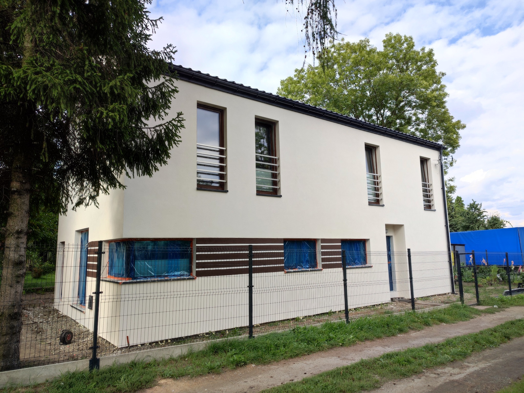 Nowoczesny, dwukondygnacyjny dom z białymi ścianami i brązowymi akcentami, okna zabezpieczone folią ochronną, widoczna metalowa siatka ogrodzeniowa i fragment zieleni.