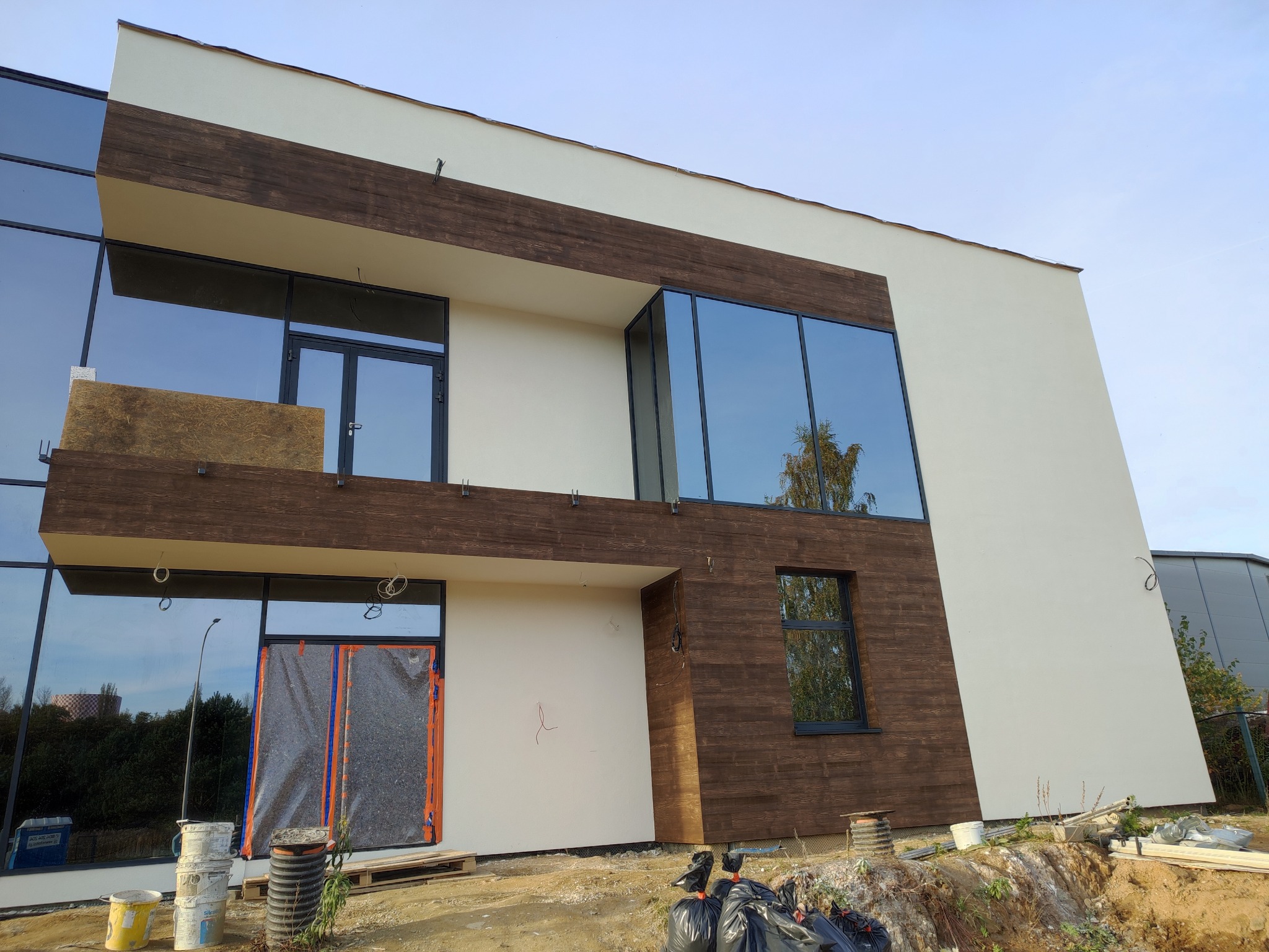 Nowoczesny dom z dużymi oknami i elewacją z elementami drewnopodobnymi w trakcie wykończenia, widoczne materiały budowlane i zabezpieczenia okien.