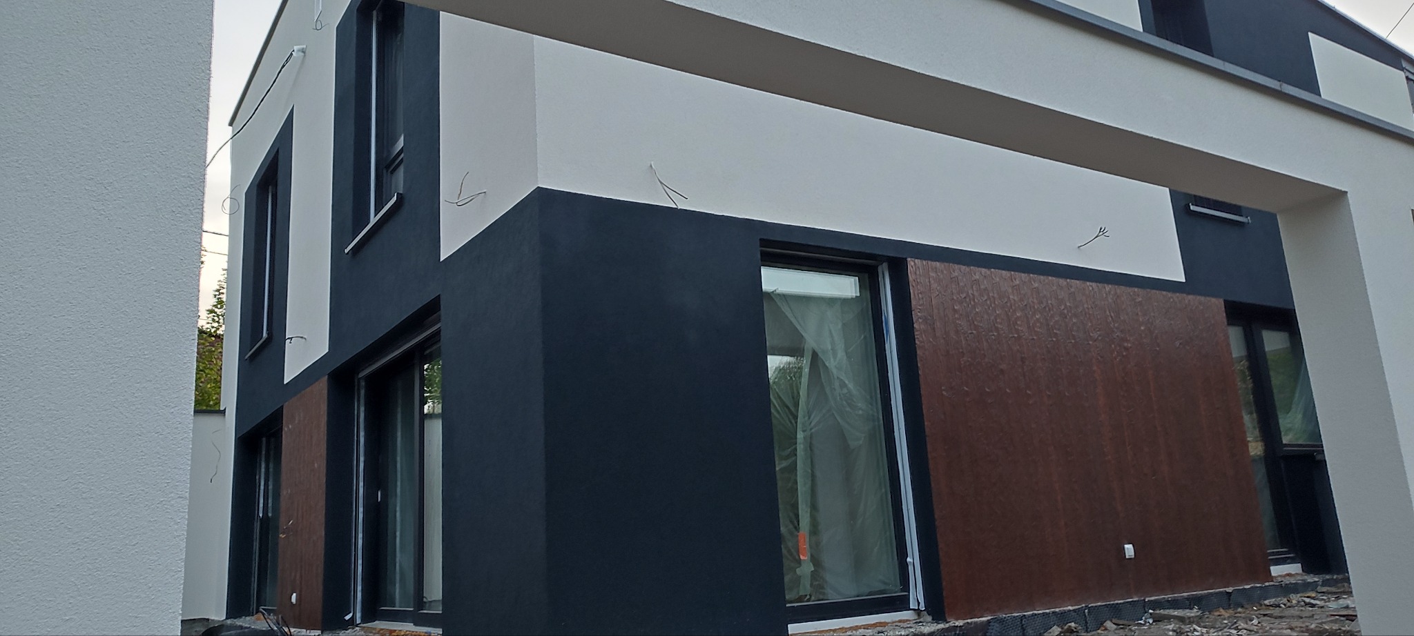 Dwukondygnacyjny dom z elewacją w kolorach bieli, szarości i brązu, z widocznymi przewodami wystającymi ze ściany i folią ochronną na oknach.