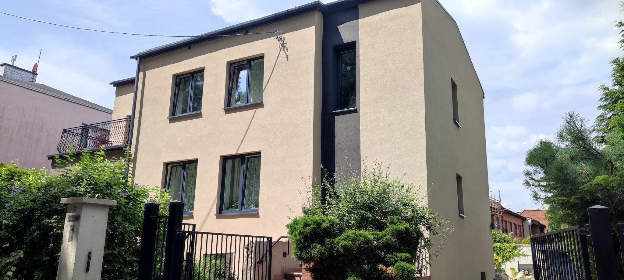 Dwukondygnacyjny dom z beżową elewacją, czarnymi ramami okiennymi i pionowym pasem w kolorze antracytowym, widoczny zza metalowego ogrodzenia i zieleni krzewów.