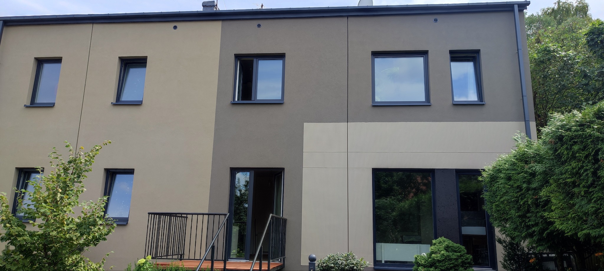 Nowoczesny dom z elewacją w dwóch odcieniach szarości i beżu, z balkonem z czarną balustradą i oknami z grafitowymi ramami.