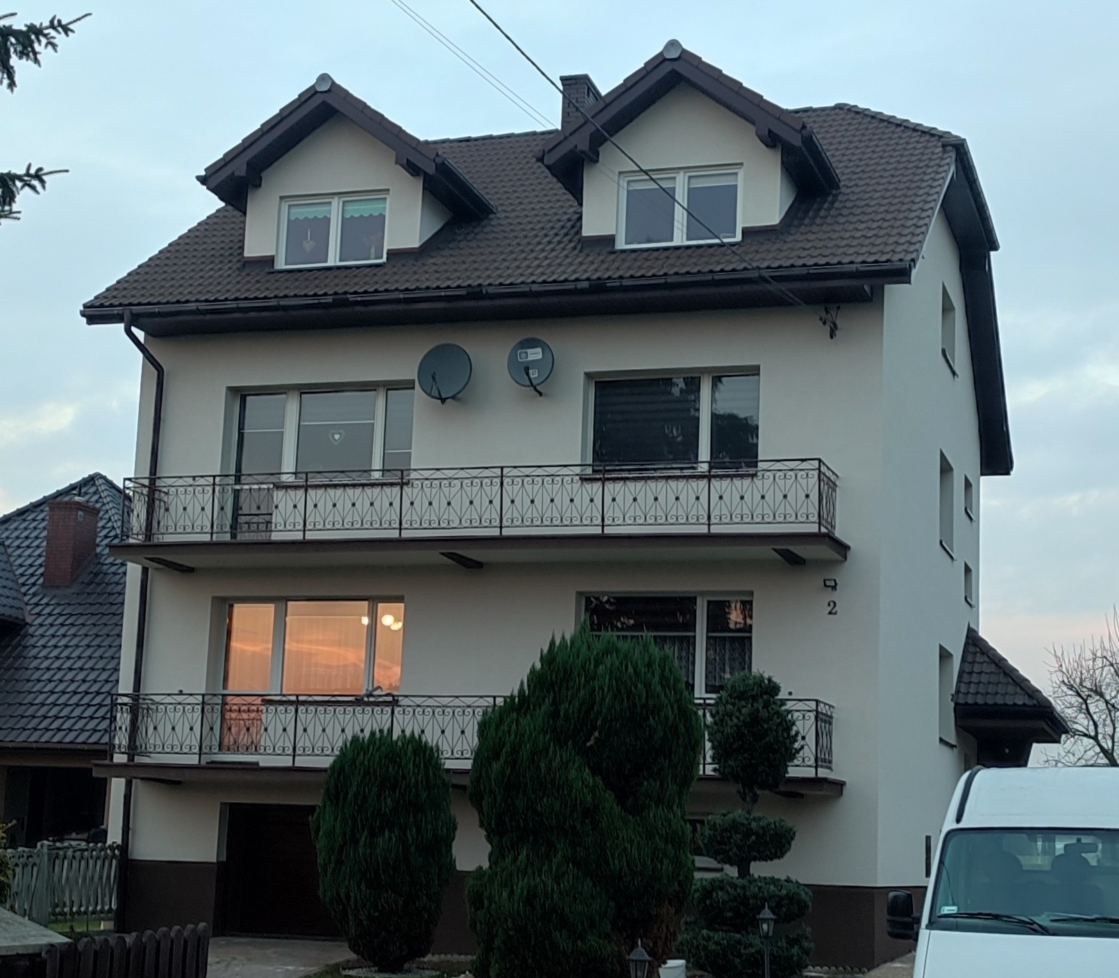 Dwupiętrowy dom z balkonami, elewacją w kolorze złamanej bieli, brązowym dachem i dwoma antenami satelitarnymi na elewacji, widok z poziomu ulicy.