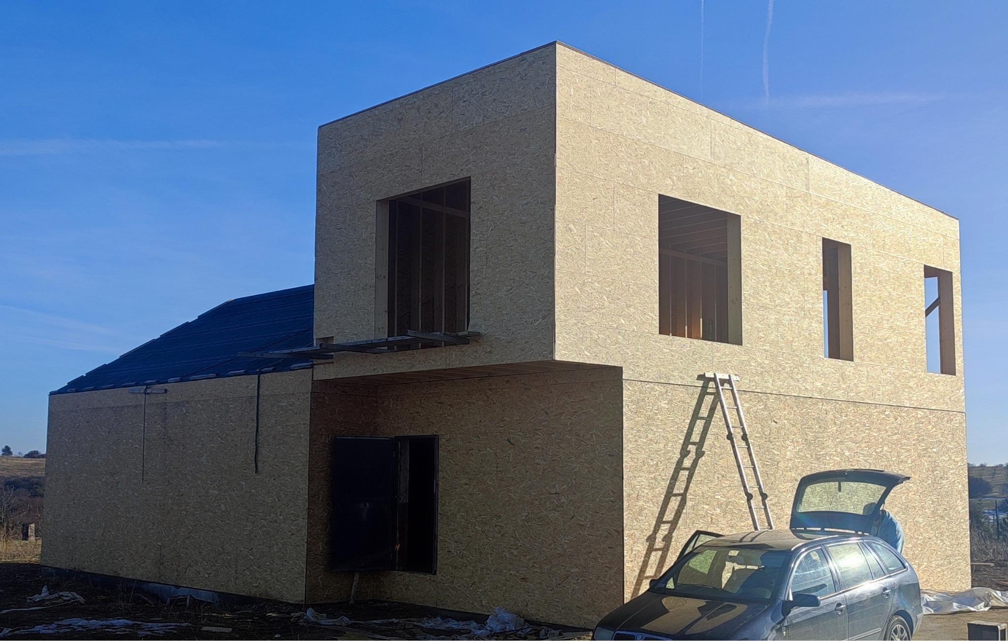 Dwukondygnacyjny dom w konstrukcji szkieletowej, z widocznym poszyciem z płyt OSB, częściowo pokryty dachem, z otworami okiennymi i drzwiami, drabina oparta o ścianę, otwarty bagażnik samochodu...