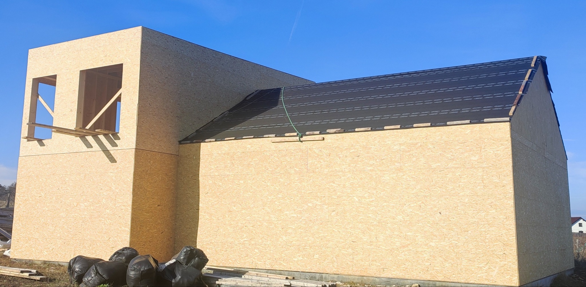 Surowy stan domu szkieletowego z płyt OSB, widoczne otwory okienne z drewnianym rusztowaniem i czarne pokrycie dachu z listwami kontrłat, worki ze śmieciami u podstawy ściany