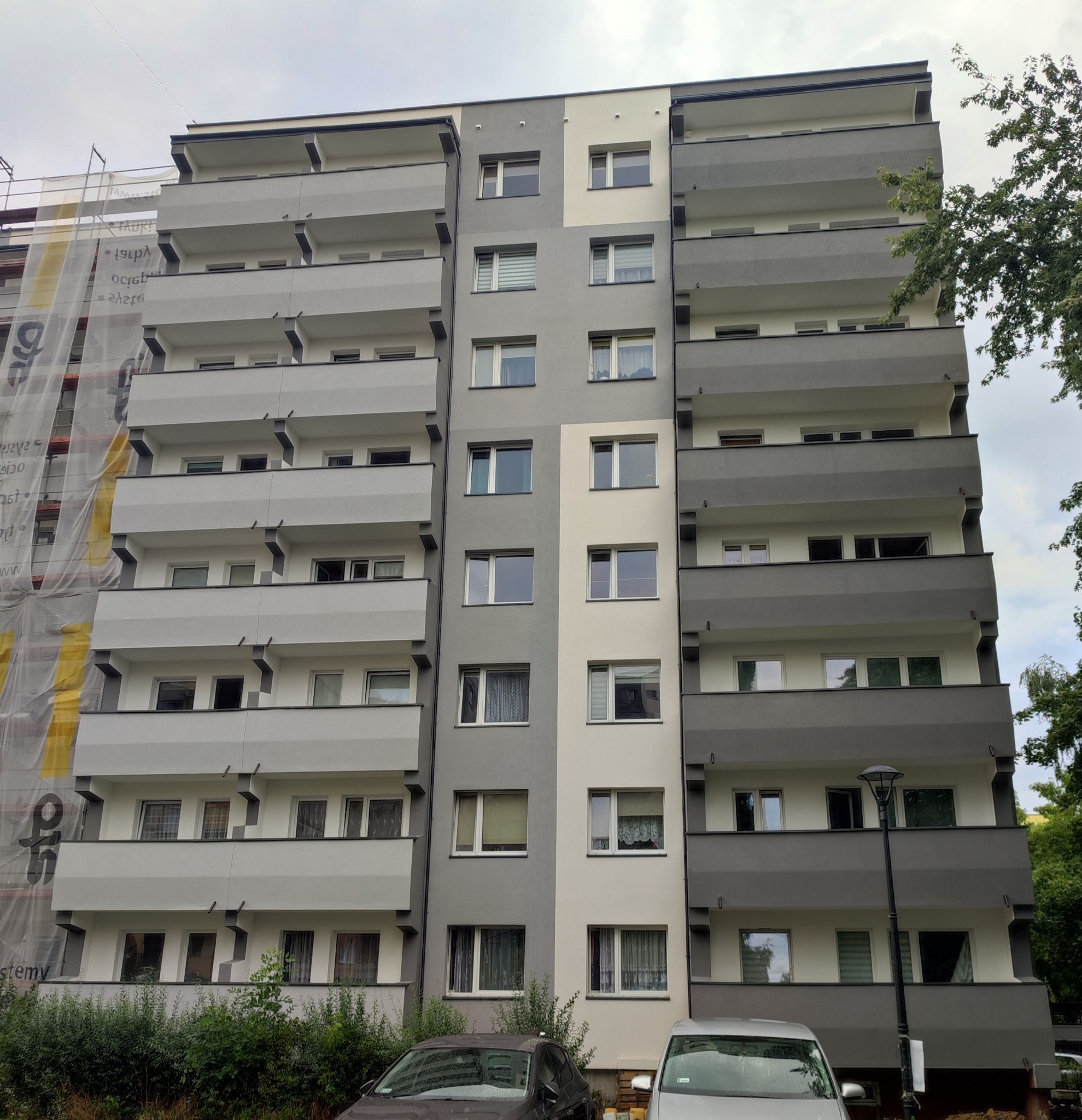Wielopiętrowy budynek mieszkalny z balkonami, pomalowany na szaro i biało, częściowo zasłonięty rusztowaniami z osłoną ochronną, z zaparkowanymi dwoma samochodami na pierwszym planie.