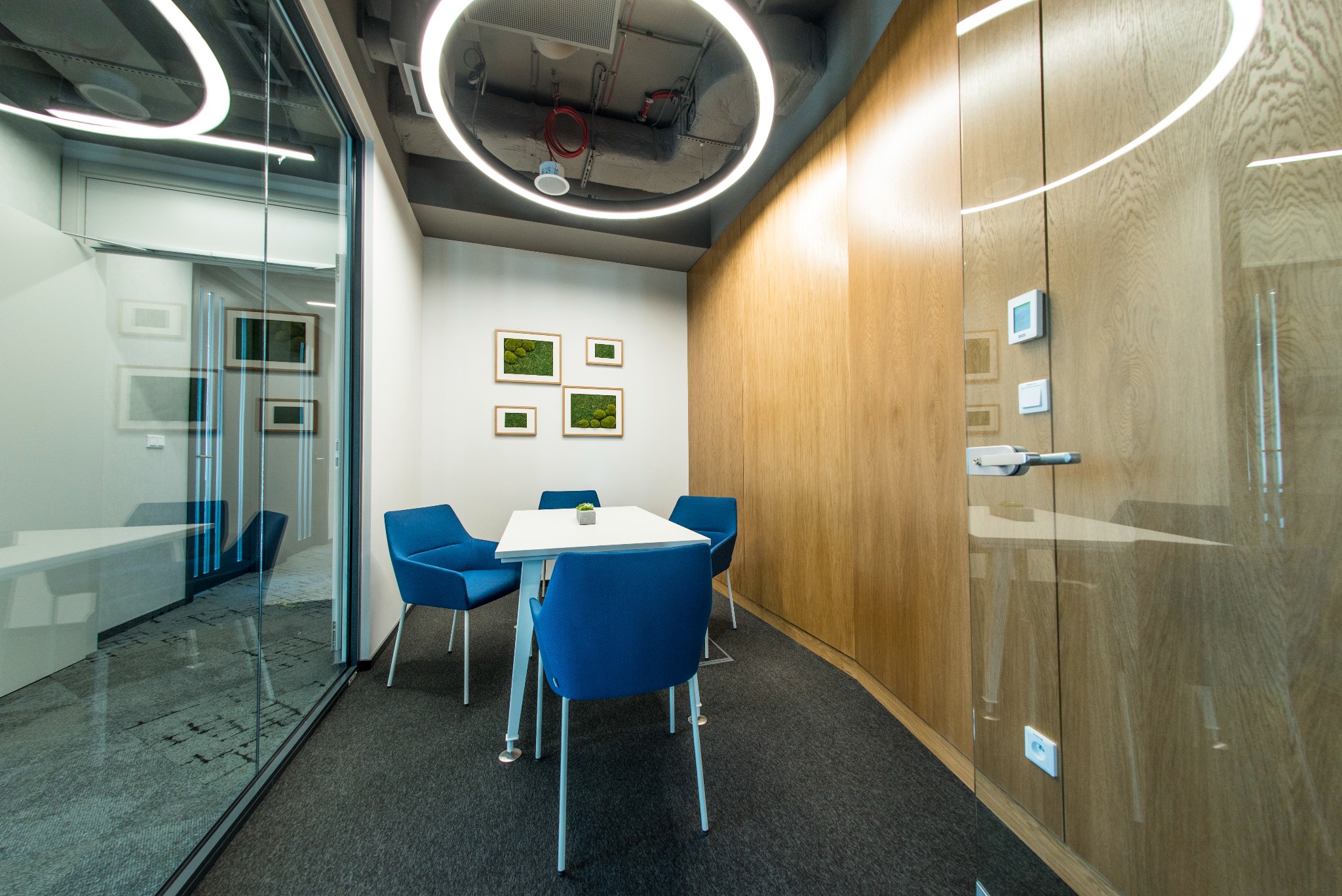 Nowoczesna sala konferencyjna z białym stołem, czterema niebieskimi krzesłami, drewnianą ścianą i okrągłym oświetleniem LED na suficie, widoczna przez szklaną ścianę.