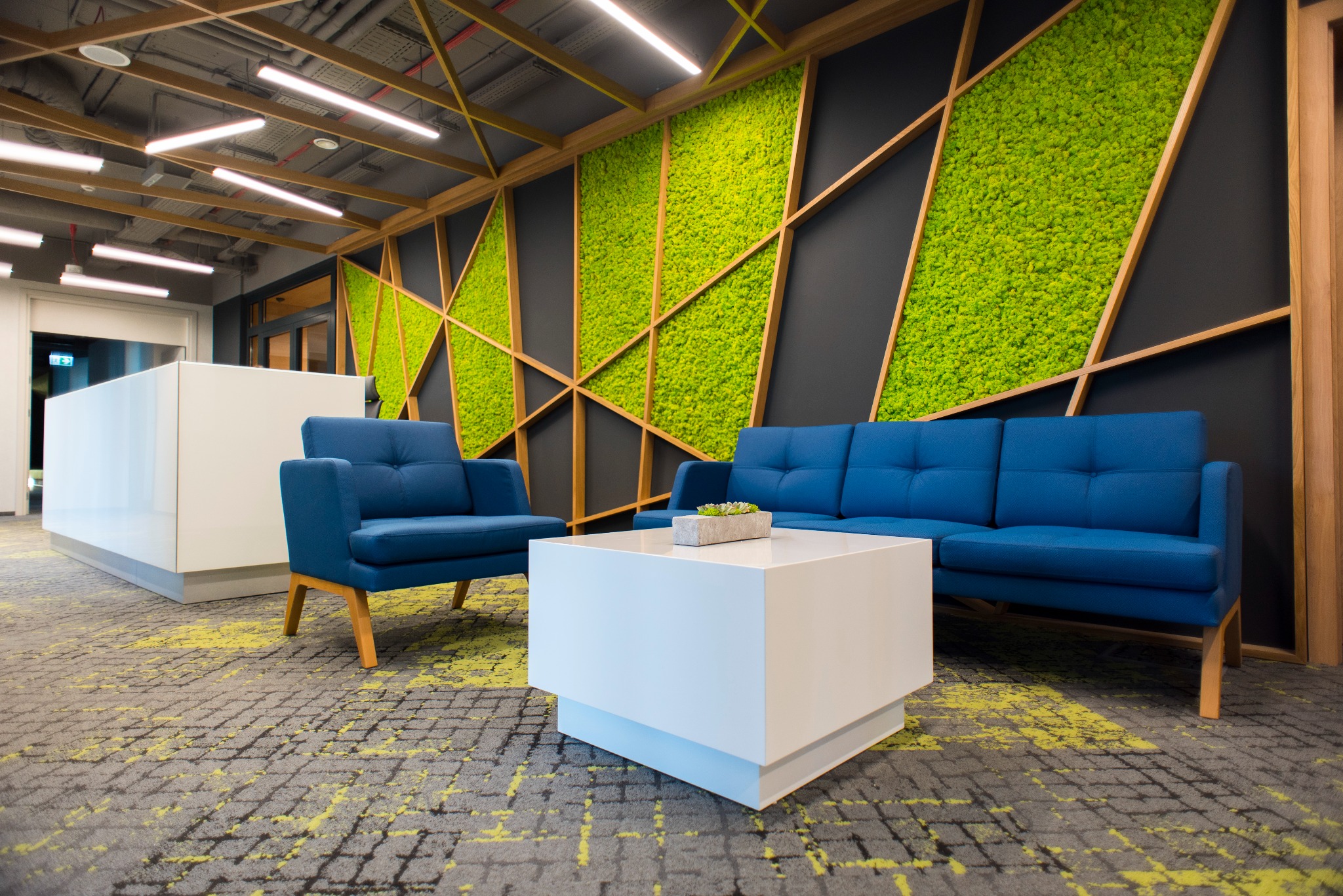Nowoczesna przestrzeń biurowa z niebieskimi sofami, białym stolikiem kawowym i ścianą z mchu w geometrycznej drewnianej ramie.