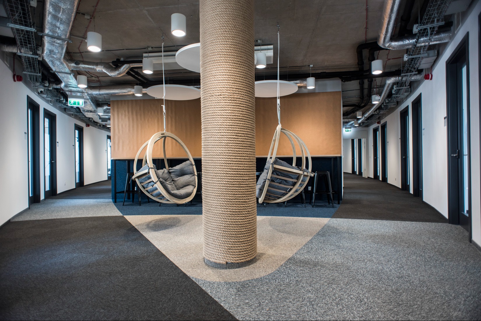 Nowoczesna przestrzeń biurowa z dwoma wiszącymi fotelami hamakowymi obok kolumny owiniętej grubym sznurem, widok na korytarz z drzwiami.