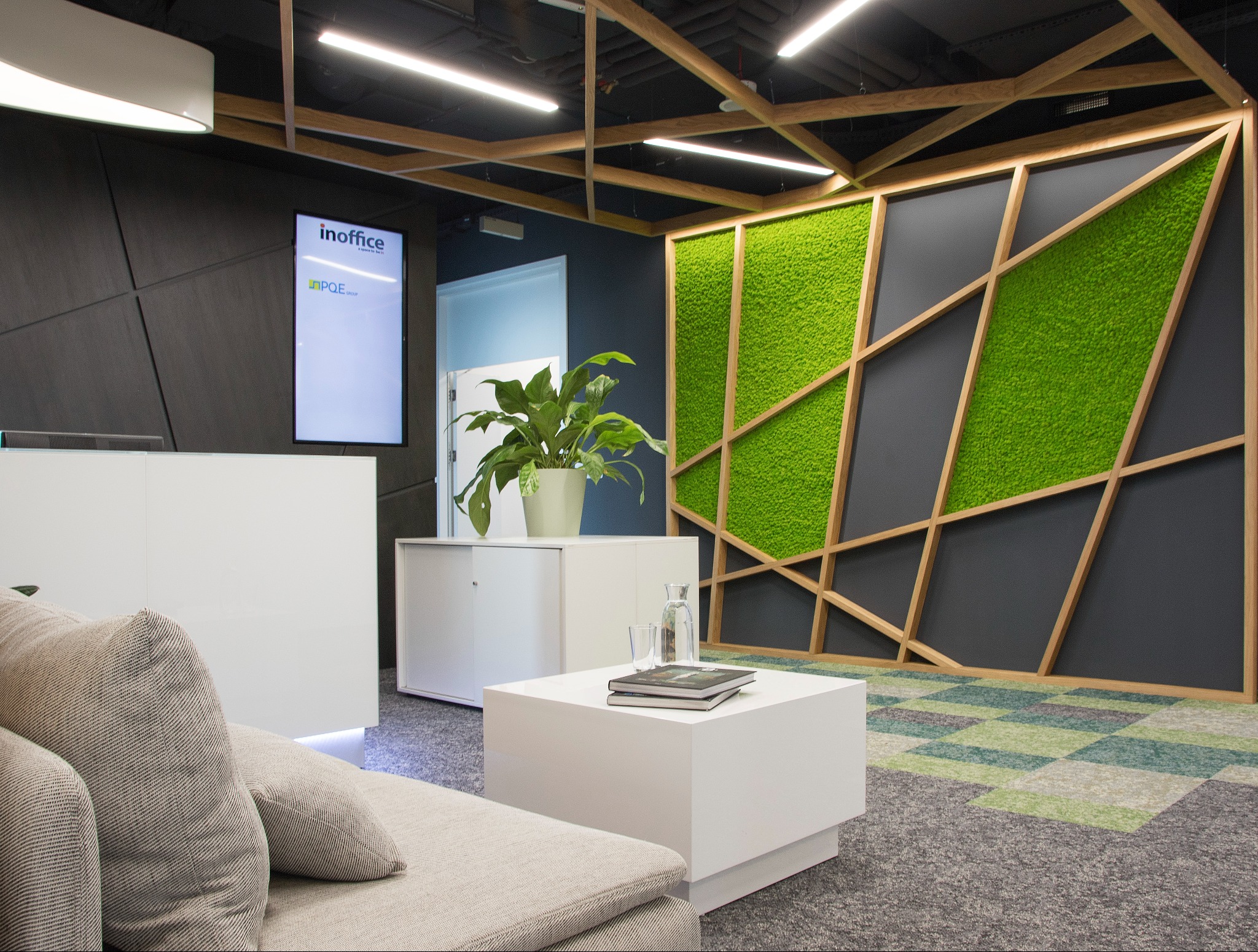 Nowoczesne wnętrze biurowe z recepcją, zieloną ścianą z mchu w drewnianej ramie, minimalistycznymi meblami i ekranem z logo firmy.