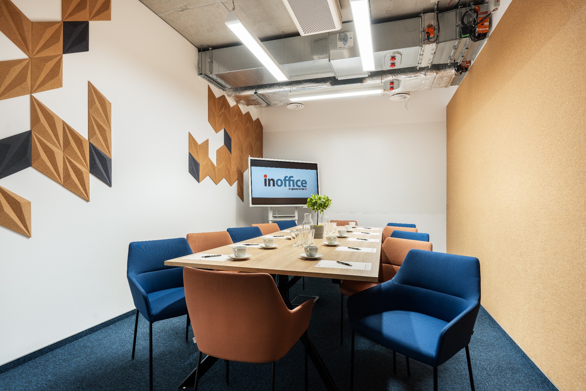 Sala konferencyjna z długim stołem, niebieskimi i pomarańczowymi krzesłami, filiżankami, notatnikami i logiem 'inoffice' na ekranie, ozdobiona geometrycznymi panelami ściennymi i korkiem.