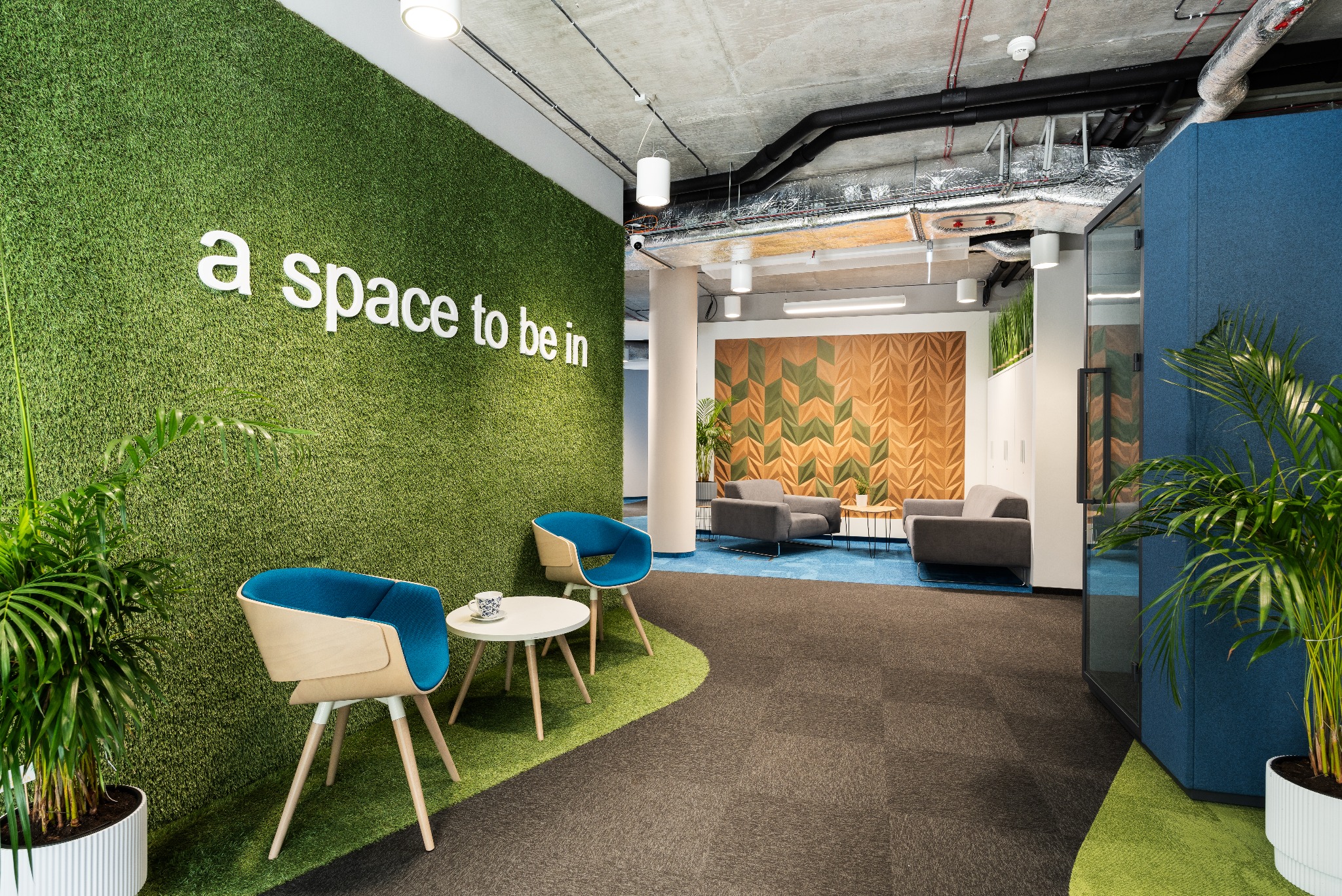 Nowoczesna przestrzeń biurowa z zieloną ścianą z napisem 'a space to be in', błękitnymi fotelami i stolikiem z filiżanką, widok na strefę relaksu z szarymi sofami i dekoracyjną ścianą.