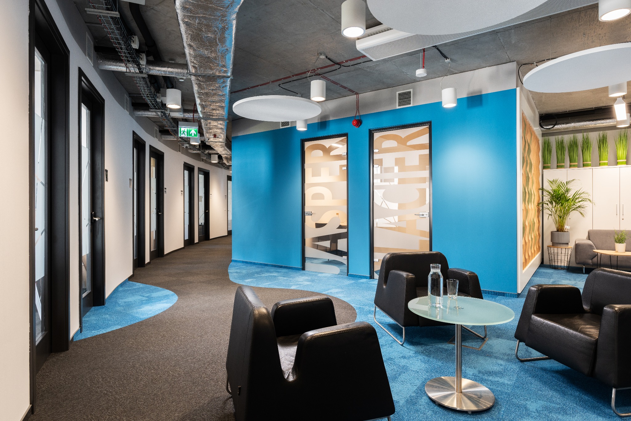Nowoczesne wnętrze biurowe z niebieską ścianą, czarnymi fotelami, szklanym stolikiem i drzwiami z nazwami firm.