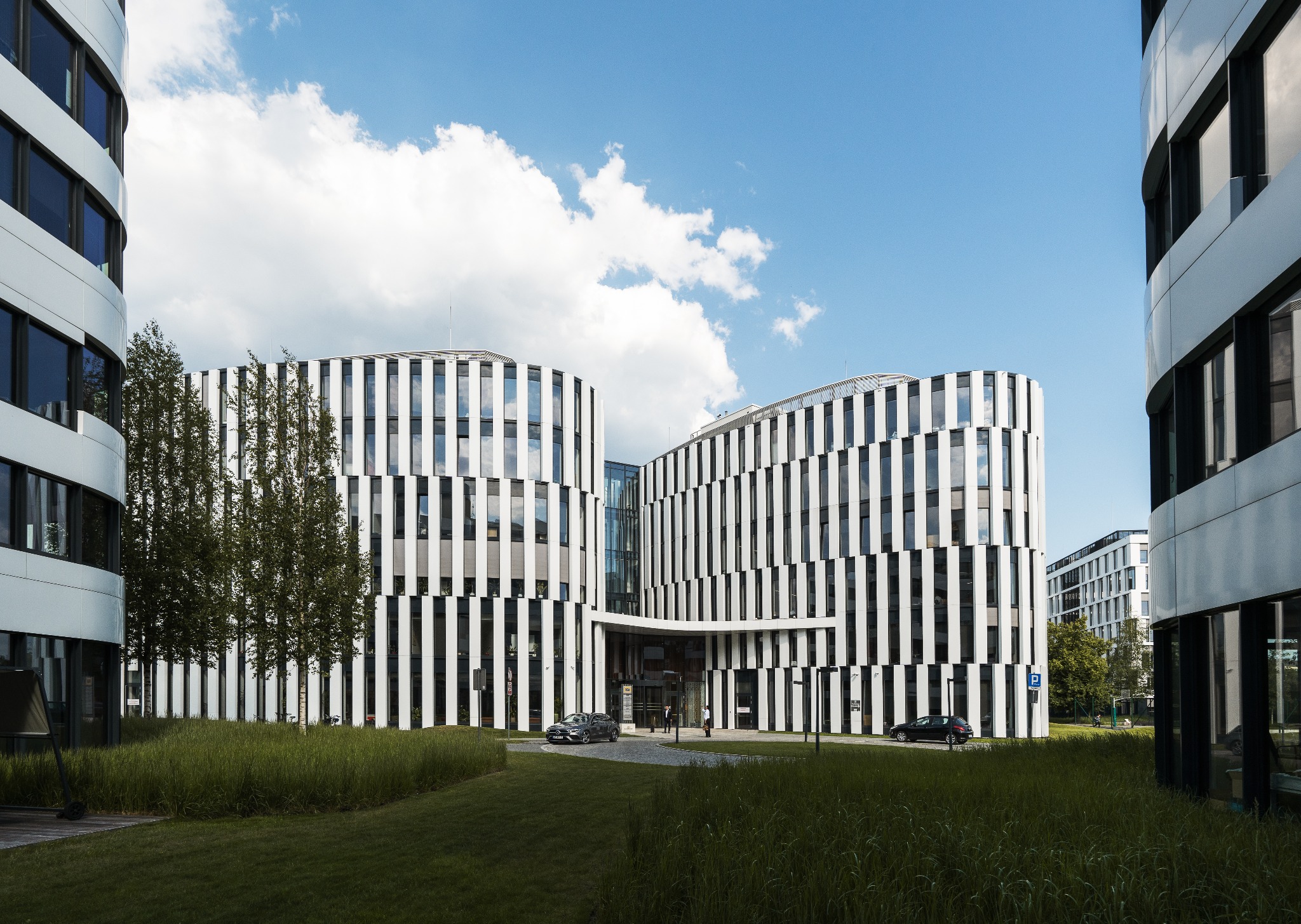 Nowoczesny budynek biurowy z czarno-białą fasadą w paski, otoczony zielonym trawnikiem i drzewami, pod błękitnym niebem z chmurami.