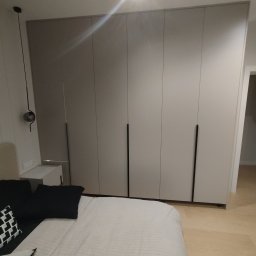 P&J Meble - Jasna szafa wnękowa z długimi, czarnymi uchwytami, umieszczona w sypialni z widocznym łóżkiem i lampką nocną. Minimalistyczny design.