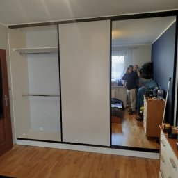 P&J Meble - Szafa wnękowa z przesuwnymi drzwiami, z lustrem odbijającym wnętrze pokoju z dwoma osobami. Widoczne półki i drążki w szafie, parkiet na podłodze.