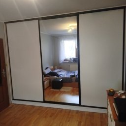 P&J Meble - Szafa przesuwna z lustrzanymi drzwiami w pokoju, odbicie wnętrza sypialni. Minimalistyczny design, jasne fronty, czarne obramowania. Parkiet na podłodze.