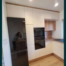 P&J Meble - Jasna kuchnia z drewnianymi akcentami: zabudowa z lodówką w czarnym połysku i piekarnikiem. Na blacie płyta grzewcza. W odbiciu widać fotografa.