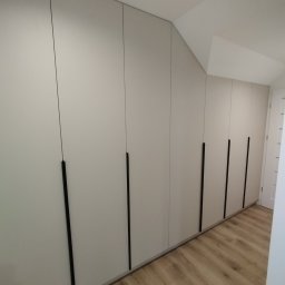 P&J Meble - Długa, zabudowana szafa z jasnymi frontami i czarnymi, pionowymi uchwytami, dopasowana do skosu sufitu, z widoczną podłogą w odcieniach drewna.