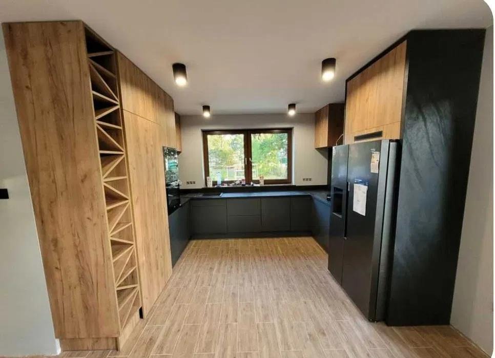 Nowoczesna kuchnia z szarymi szafkami, drewnianymi elementami i regałem na wino z trójkątnymi przegródkami, oświetlona punktowymi lampami sufitowymi i naturalnym światłem z okna.