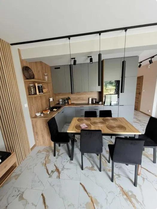 Nowoczesna kuchnia z szarymi szafkami i drewnianymi akcentami, oświetlenie szynowe, stół z czarnymi krzesłami na marmurowej podłodze.