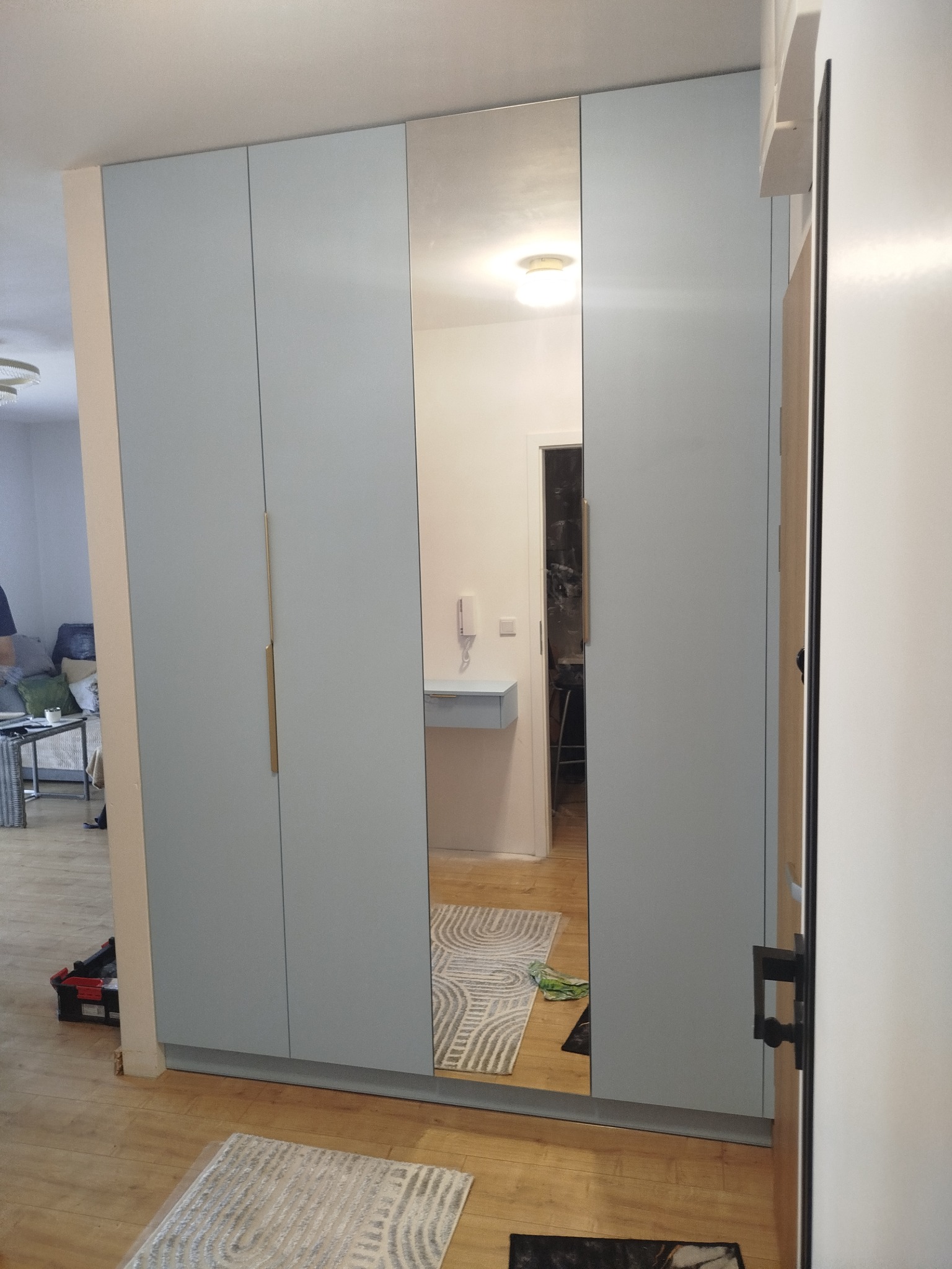 Jasnoszara szafa wnękowa z lustrem, odbijająca wnętrze mieszkania z dywanem w geometryczne wzory. Złote uchwyty dodają elegancji. Widoczna skrzynka z narzędziami.