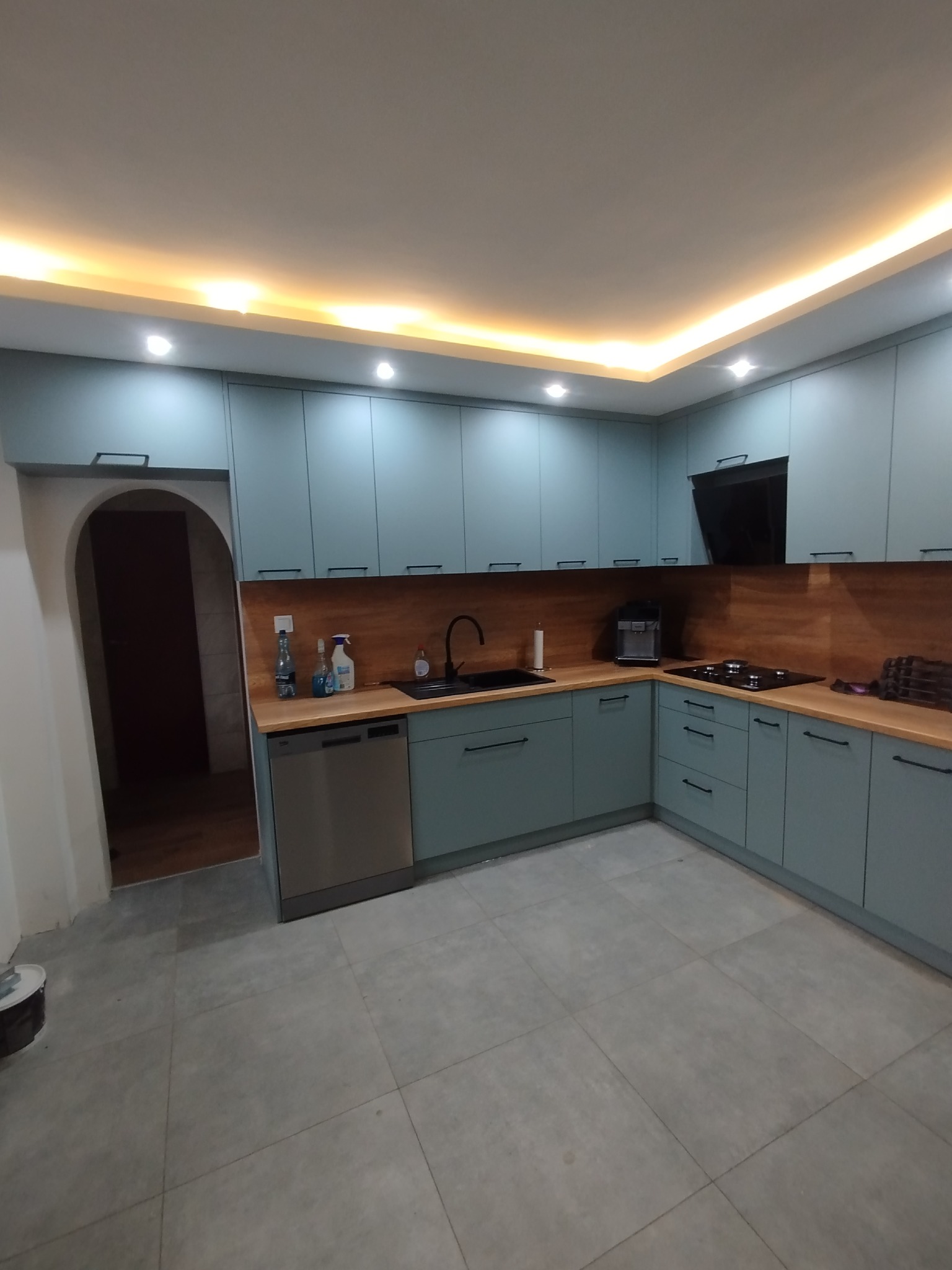 Kompletna kuchnia w odcieniach błękitu z drewnianym blatem i czarnymi uchwytami, oświetlona listwą LED pod sufitem. Widok na zlew, płytę grzewczą i sprzęt AGD.