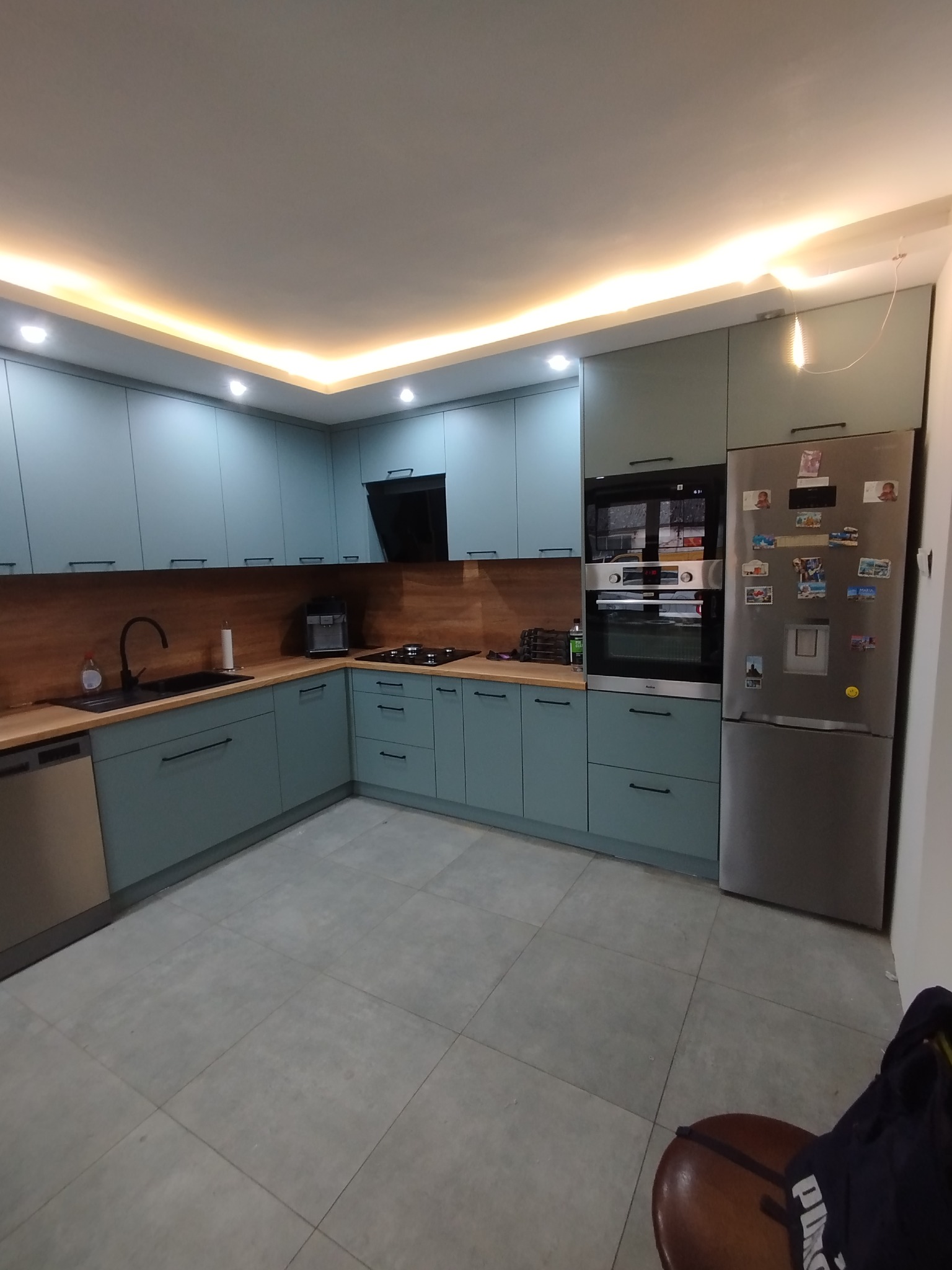 Narożna kuchnia z turkusowymi szafkami i drewnianym blatem, oświetlona ledami pod sufitem. Srebrna lodówka z magnesami. Minimalistyczny design.